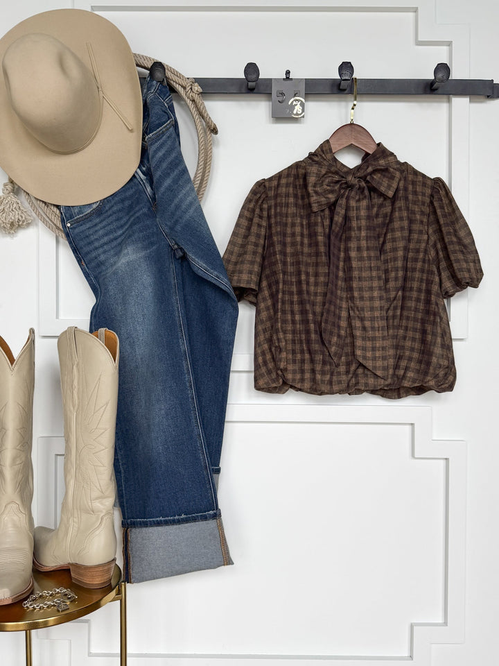 Western outfit: beige cowboy hat, blue jeans, cream cowboy boots, and the Savannah Sevens western life{&}style Ellendale Chocolate Gingham Bubble Hem Top with a bow tie, all displayed against a white wall on hooks and a gold stool.