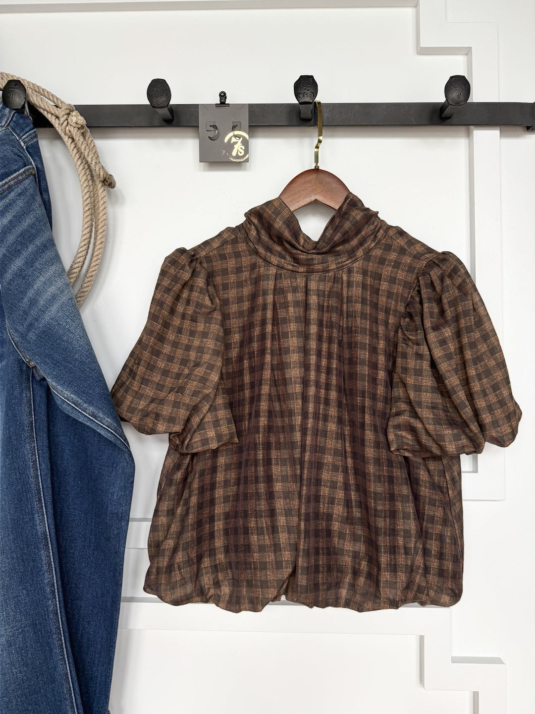 The Ellendale Chocolate Gingham Bubble Hem Top by Savannah Sevens western life{&}style, featuring puffed short sleeves, hangs on a black wall hook beside blue jeans draped over the same hook, with a white textured wall in the background.