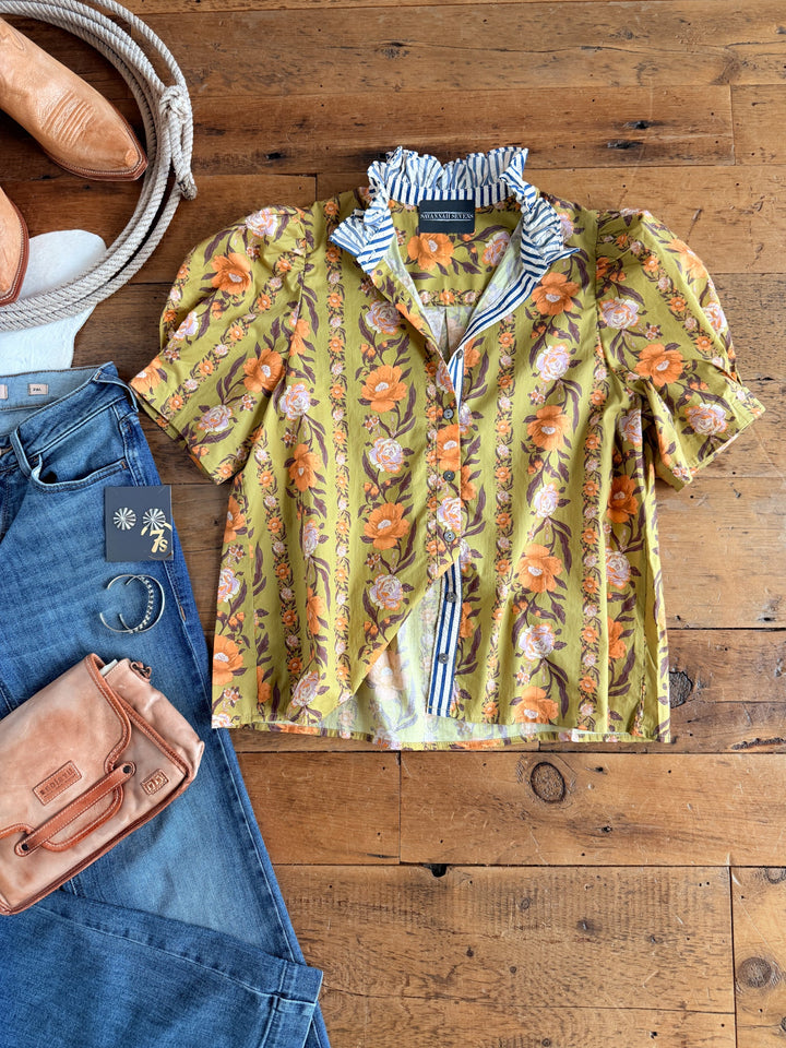 The Ellington Chartreuse Floral Top from Savannah Sevens western life{&}style is styled with blue jeans, a tan purse, silver hoop earrings, and cowboy boots, all laid out on a wooden floor.