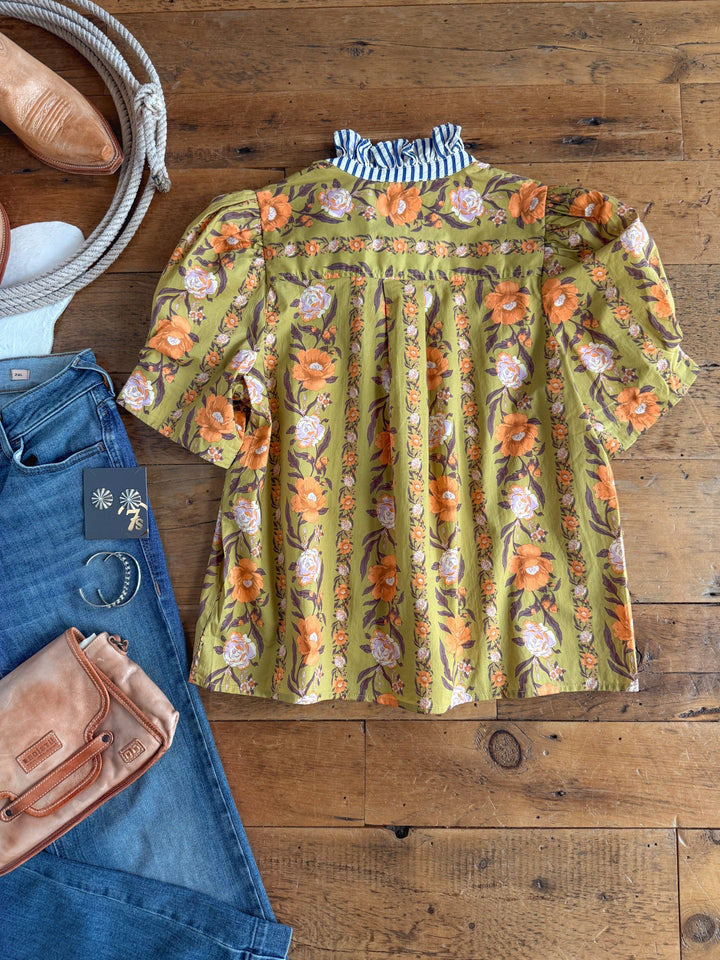 The Ellington Chartreuse Floral Top by Savannah Sevens western life{&}style—a button-up blouse with puff sleeves and a striped ruffle high neck collar—is laid flat on a wooden floor, surrounded by blue jeans, a tan purse, brown shoes, earrings, and a white rope.