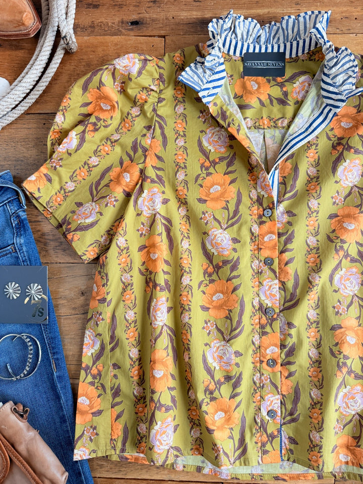 The Ellington Chartreuse Floral Top by Savannah Sevens western life{&}style, featuring yellow and orange florals, puff sleeves, and a blue striped ruffle high neck collar, is displayed on a wooden surface next to blue jeans and brown leather accessories.