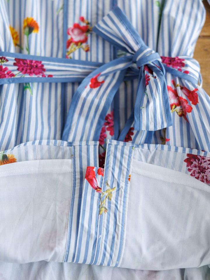 A close-up of the Ellinwood Floral Striped Cotton Dress by Savannah Sevens western life{&}style shows its blue and white stripes, colorful floral patterns, matching sash bow at the waist, and a visible pocket edge.