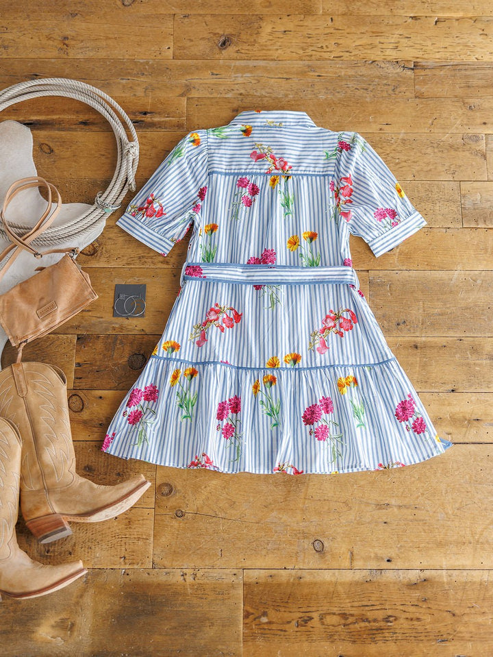 The Ellinwood Floral Striped Cotton Dress by Savannah Sevens western life{&}style, featuring blue and white stripes with pink, yellow, and red floral prints, is styled with tan cowboy boots, a brown purse, and silver hoop earrings on a wooden floor.
