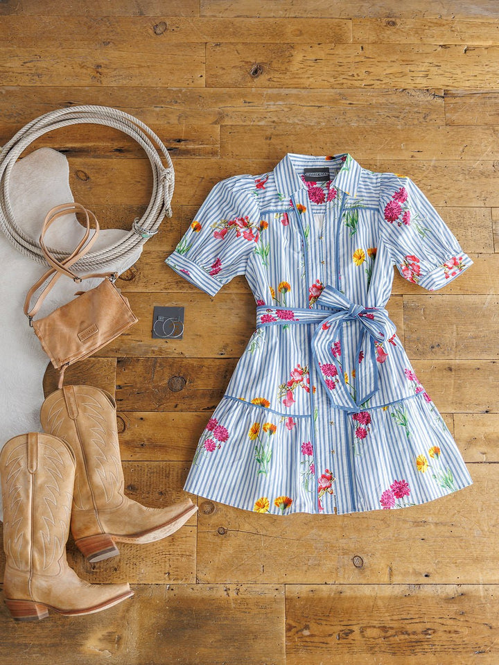 The Ellinwood Floral Striped Cotton Dress by Savannah Sevens western life{&}style is displayed on a wooden floor with tan cowboy boots, a brown crossbody bag, silver jewelry, and a rope beside it.