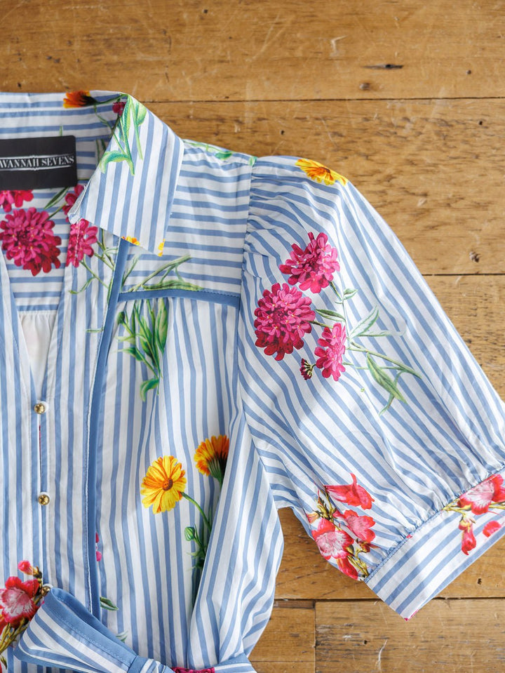 The Ellinwood Floral Striped Cotton Dress by Savannah Sevens western life{&}style features blue and white stripes, a collar, short puff sleeves, colorful floral patterns, and front buttons. Displayed on a wooden surface.