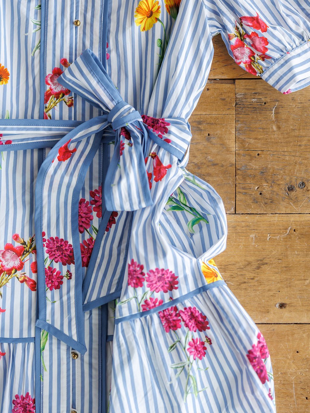 The Ellinwood Floral Striped Cotton Dress by Savannah Sevens western life{&}style features blue-and-white stripes, vibrant red, pink, and yellow floral prints, a tied waist sash, and short sleeves—perfect for adding charm to your summer wardrobe.