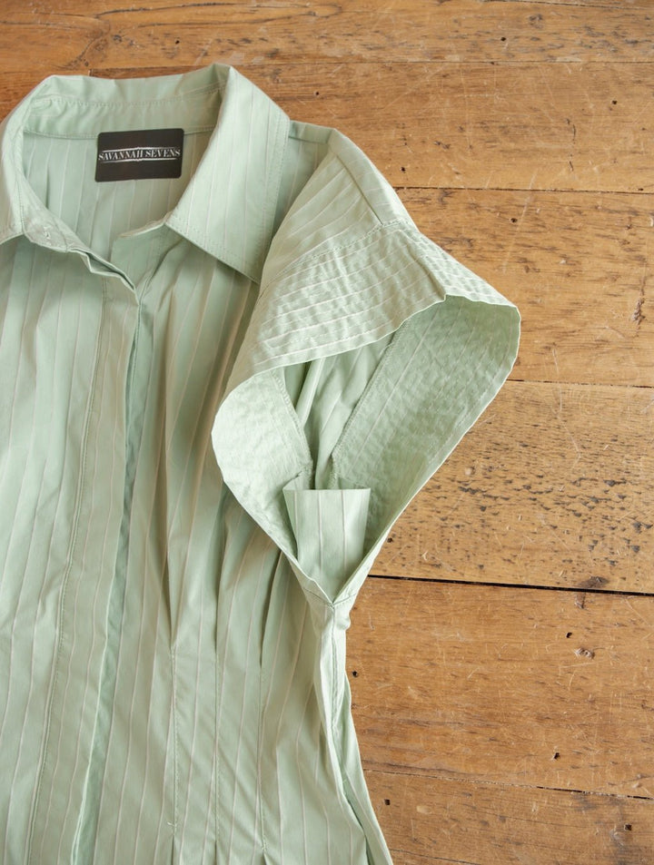 The Elmira Sage Pinstripe Button Up Dress by Savannah Sevens western life{&}style, featuring a light green hue and vertical pinstripes, is displayed flat on a wooden surface with the label and part of one sleeve visible, bringing a fresh update to classic short-sleeve button-up dress styles.