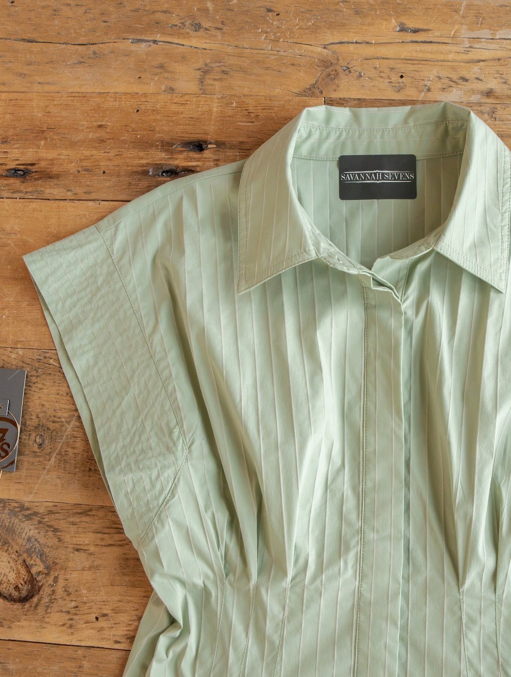 Elmira Sage Pinstripe Button Up Dress by Savannah Sevens western life{&}style, featuring light green fabric with vertical pinstripes and short sleeves, is laid flat on a wooden surface. The label inside the collar displays the brand name.