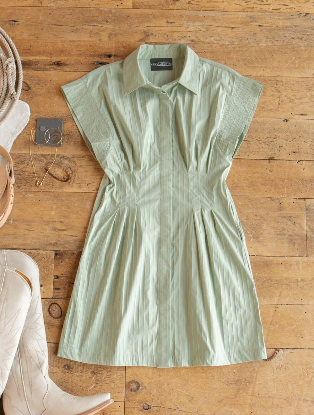 The Elmira Sage Pinstripe Button Up Dress from Savannah Sevens western life{&}style, featuring short sleeves, is laid flat on a wooden floor. Beige cowboy boots and gold jewelry are displayed nearby, with a rope partially visible on the left side.