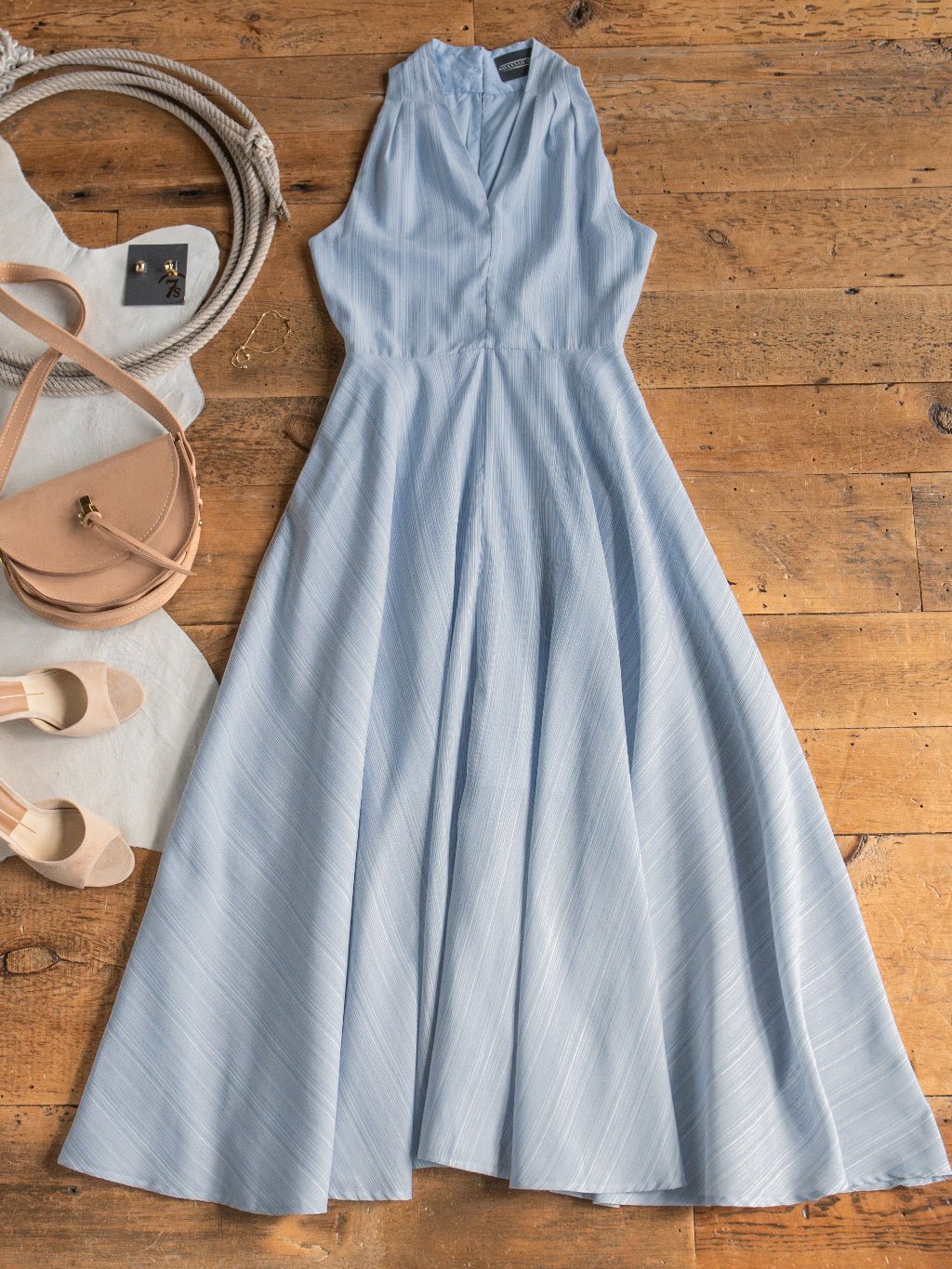 The Elnora Micro Striped Halter Neck Dress by Savannah Sevens western life{&}style is displayed on a wooden floor, paired with a beige crossbody bag, matching shoes, white fabric, rope, and a small jewelry.