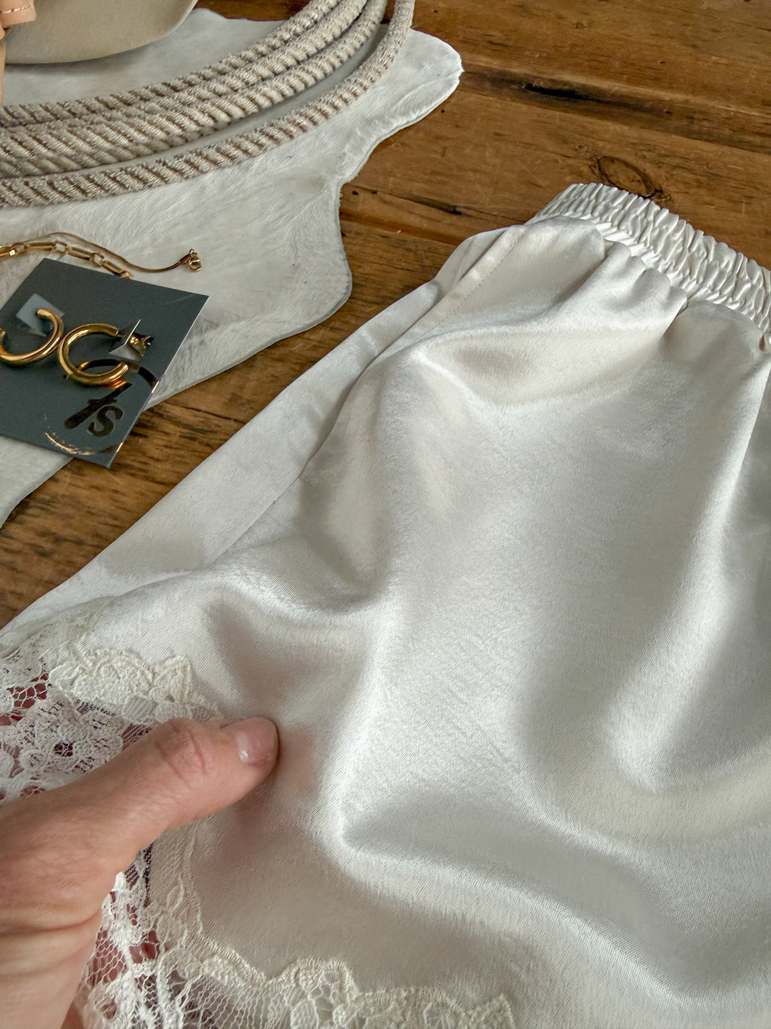 A hand holds a silky white slip with lace trim on a wooden table, next to gold hoop earrings, a necklace, and a light-colored purse. Nearby, the Elyria Satin Lace Trim Shorts from Savannah Sevens western life{&}style—with their off-white satin fabric and delicate lace hem—add an elegant touch to the scene.