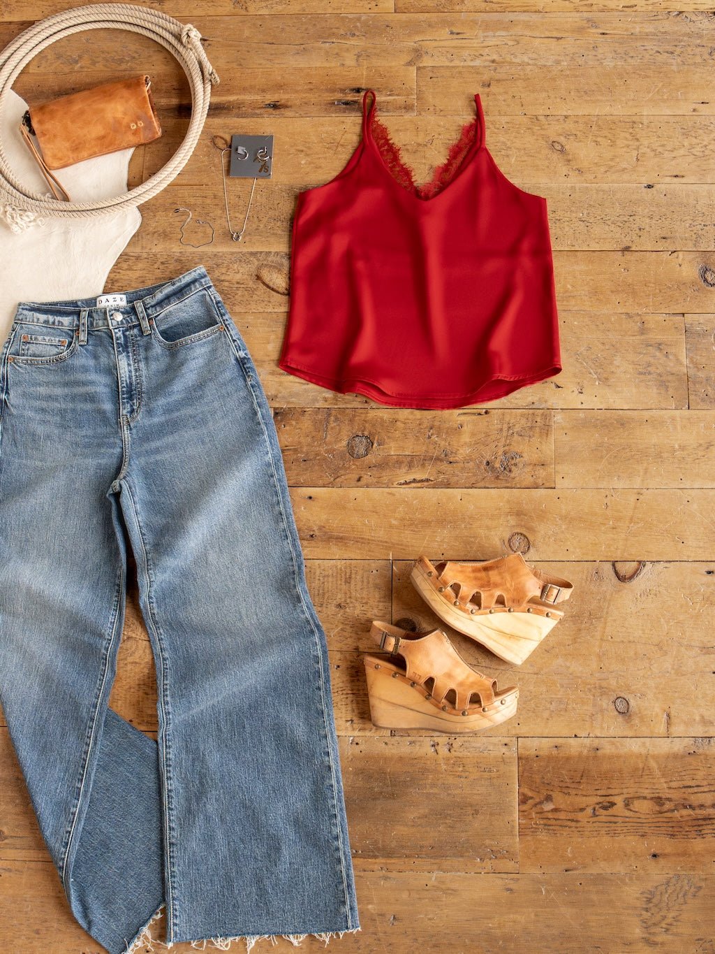 Flat lay of the Savannah Sevens western life{&}style Enderlin Red Lace Trim Satin Tank, blue jeans, tan wooden platform sandals, a cream bag with a tan pouch, and silver earrings on a wooden floor.