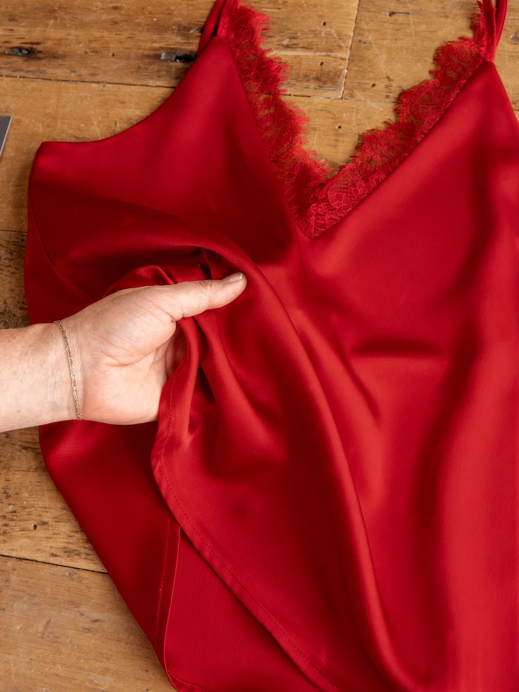A hand touches the smooth fabric of the Savannah Sevens western life{&}style Enderlin Red Lace Trim Satin Tank, with its lace-trimmed v-neck, as it rests on a wooden surface.