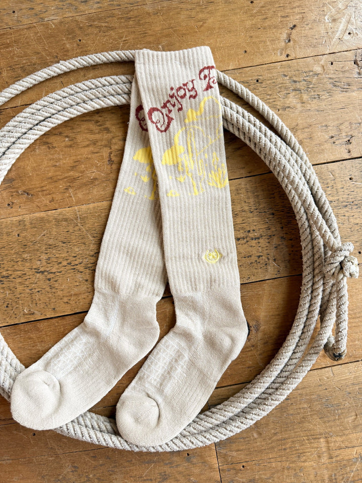 A pair of Enjoy the Ride Crew Socks from Savannah Sevens western life{&}style, featuring cream coloring with yellow and red designs, is displayed on a coiled rope atop a wooden floor.