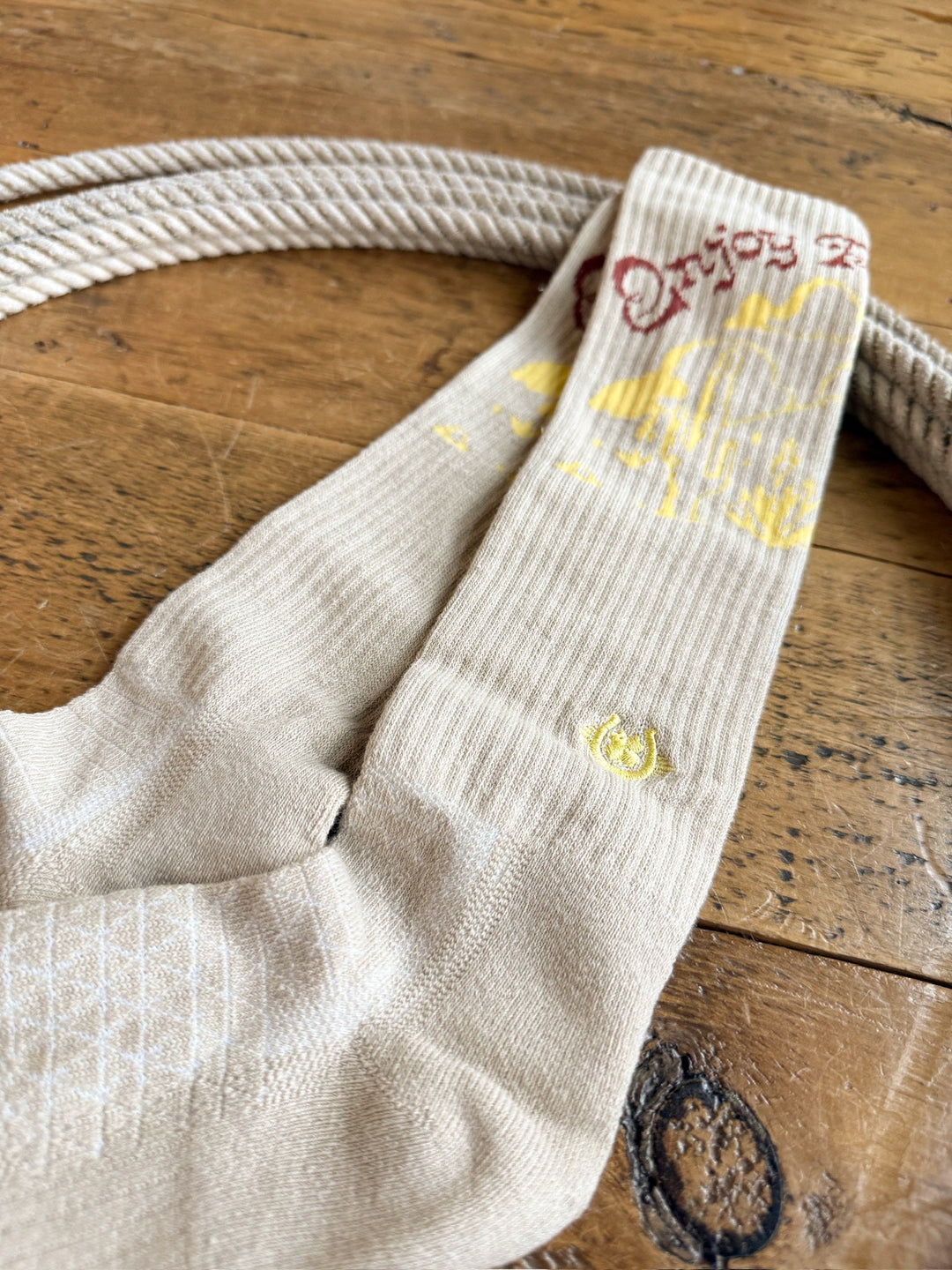 The “Enjoy the Ride Crew Socks” from Savannah Sevens western life{&}style, featuring yellow and red designs on a beige background, are displayed on a wooden surface beside a coiled rope.