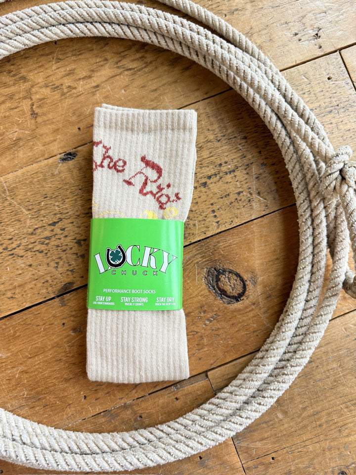 A pair of beige “Enjoy the Ride Crew Socks” by Savannah Sevens western life{&}style, featuring red text, displayed inside a coiled rope on a wooden surface.