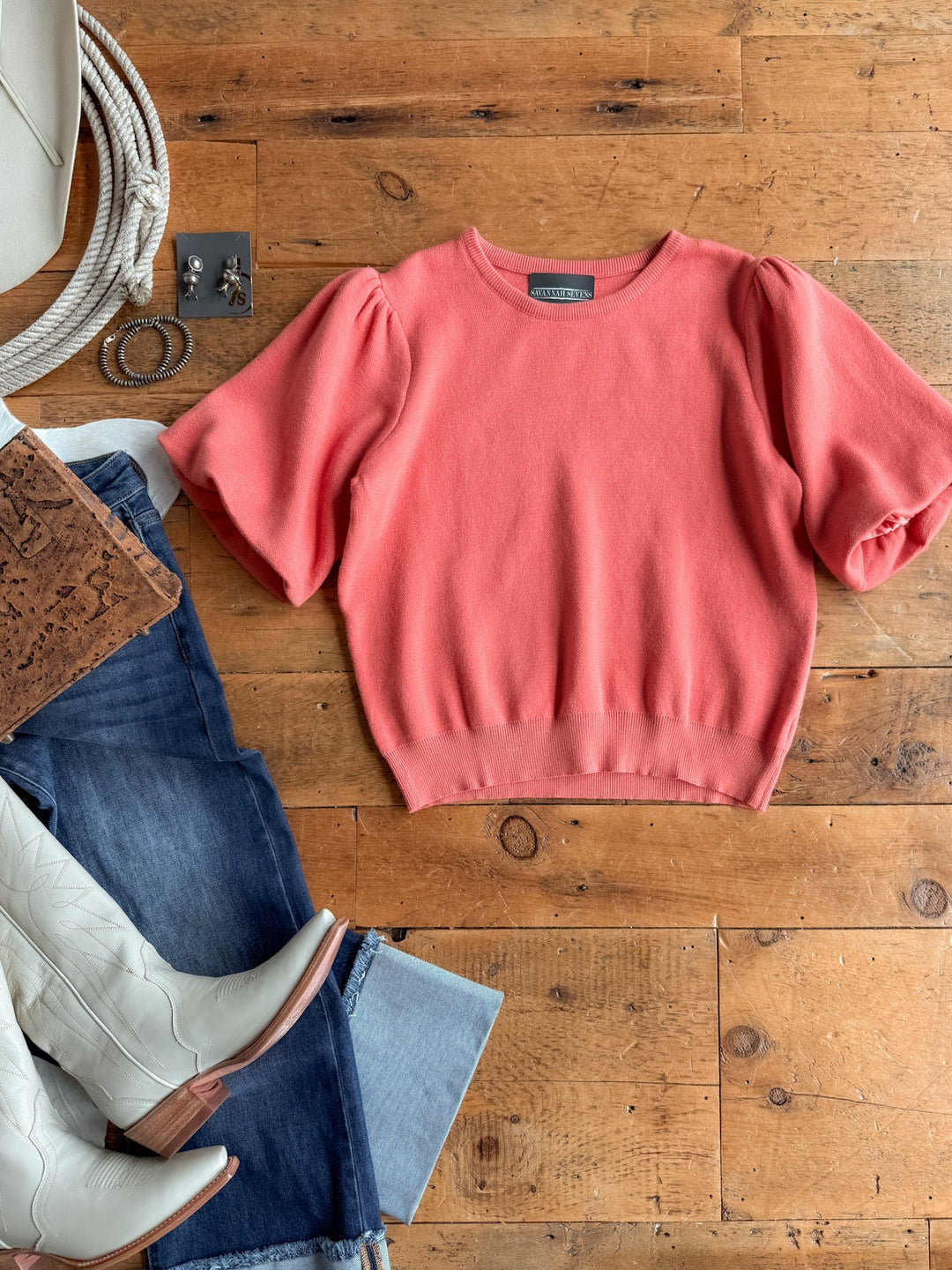 The Estacada Salmon Knit Top by Savannah Sevens western life{&}style is laid on a wooden floor with blue jeans, white cowboy boots, a white purse, silver earrings, a rope, and a brown notebook arranged nearby.