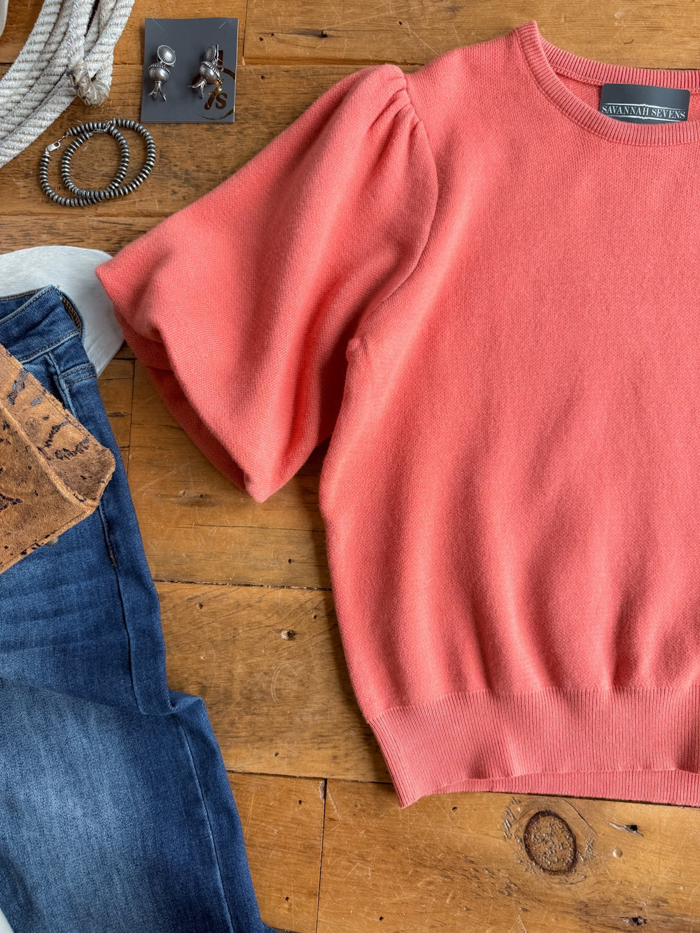 The Estacada Salmon Knit Top by Savannah Sevens western life{&}style, featuring short statement sleeves, is displayed on a wooden surface alongside blue jeans, a brown clutch, a rope necklace, stud earrings, and a silver bracelet.