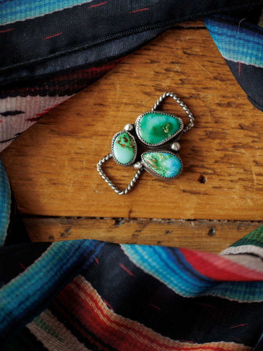 The Etowah Scarf Pendant by Savannah Sevens western life{&}style rests on wood, framed by colorful woven fabric with blue, red, black, and white patterns.