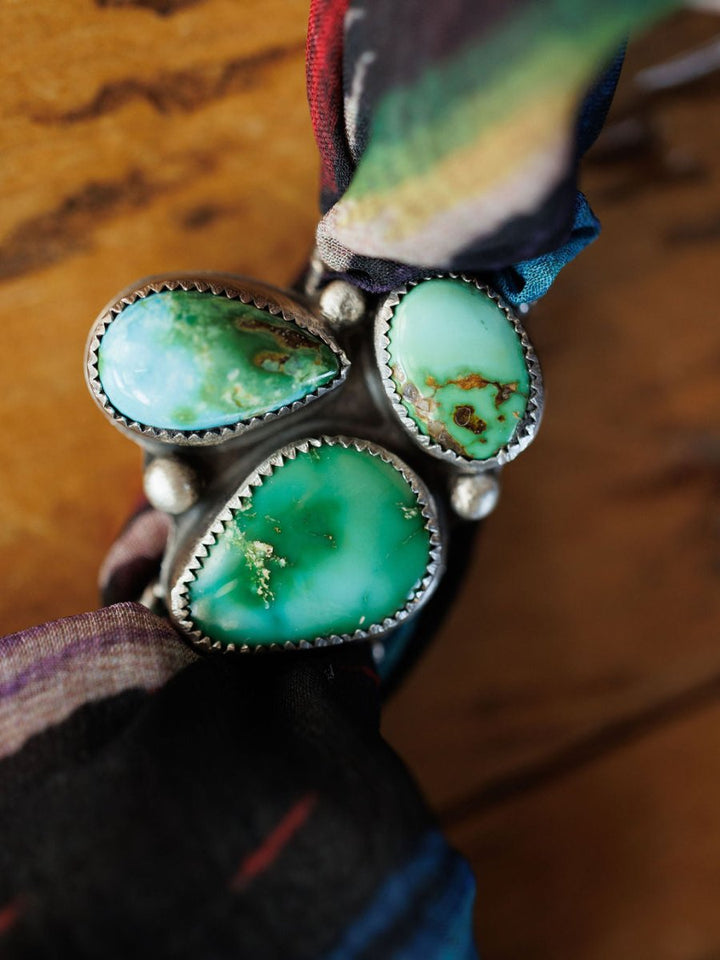 A close-up of the Etowah Scarf Pendant by Savannah Sevens western life{&}style, crafted in sterling silver with three uniquely shaped green Kingman turquoise stones, shown on a colorful fabric against a wooden background.