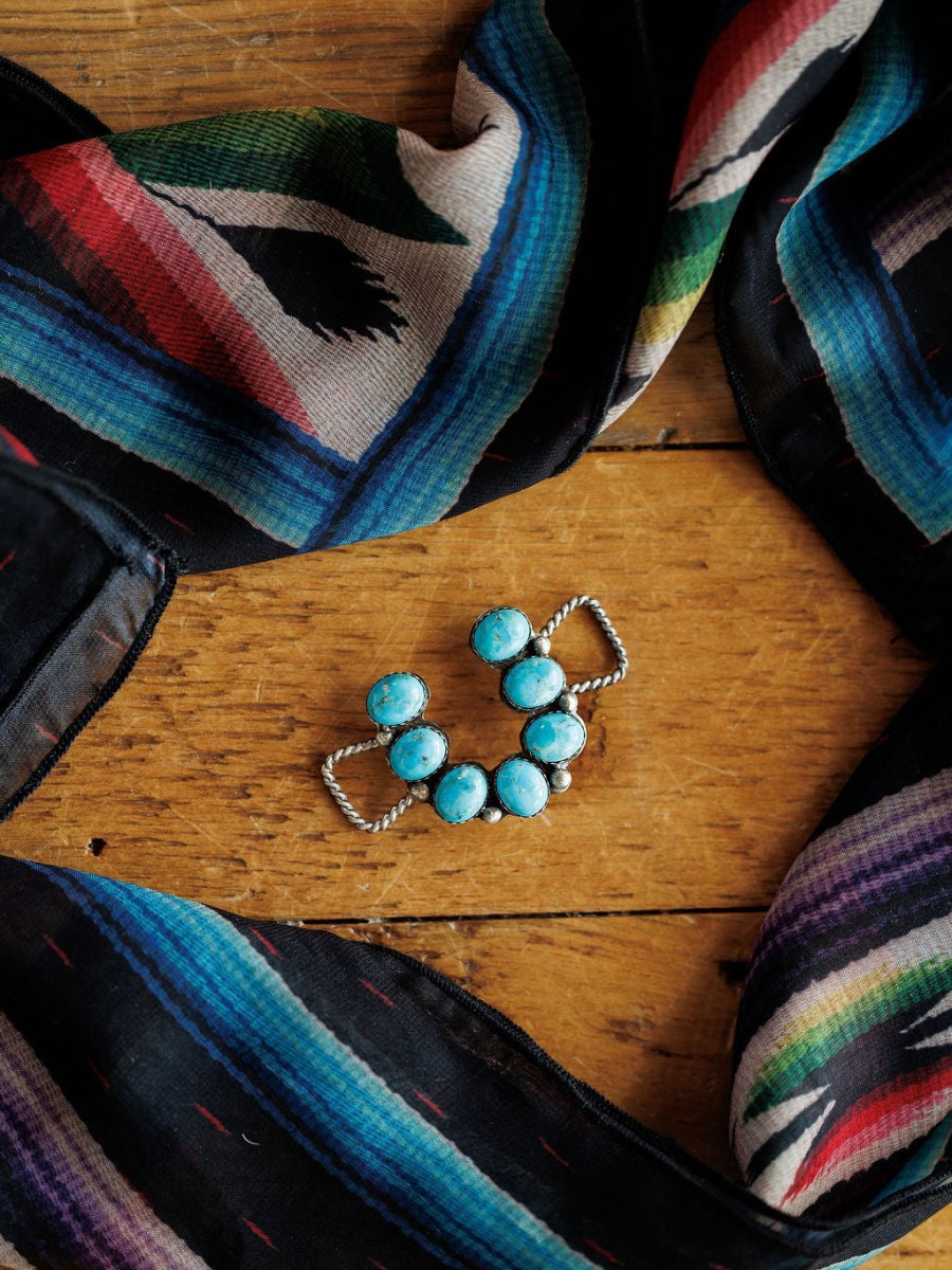 The Etowah Scarf Pendant by Savannah Sevens western life{&}style sits on wood, framed by a colorful scarf featuring blue, green, red, and purple stripes with geometric designs.