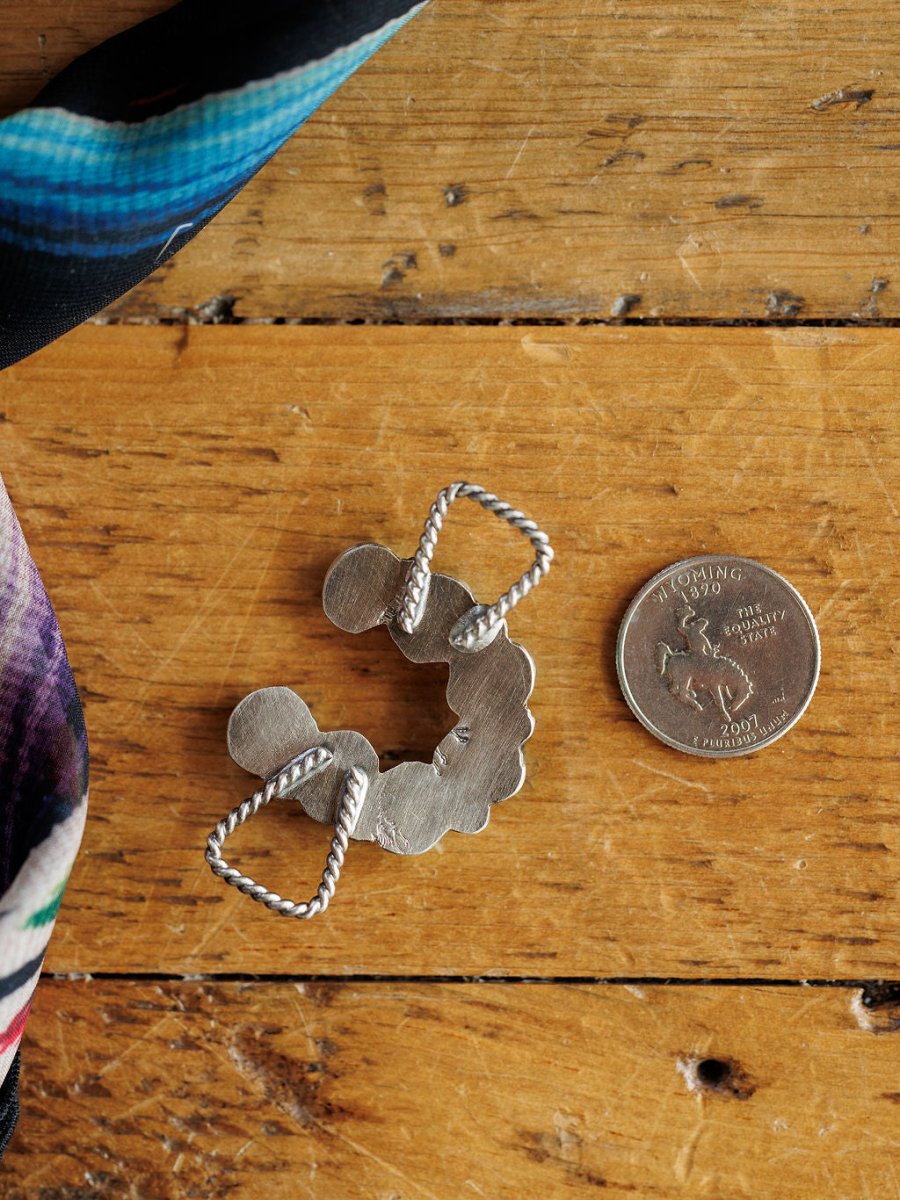 The Etowah Scarf Pendant by Savannah Sevens western life{&}style, featuring a twisted rope design in sterling silver, rests on wood beside a Wyoming quarter for scale. A colorful scarf peeks from the corner, ideal for pairing as a turquoise scarf pendant.