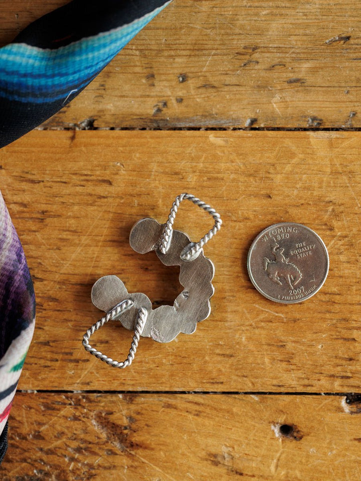 The Etowah Scarf Pendant by Savannah Sevens western life{&}style, featuring a twisted rope design in sterling silver, rests on wood beside a Wyoming quarter for scale. A colorful scarf peeks from the corner, ideal for pairing as a turquoise scarf pendant.