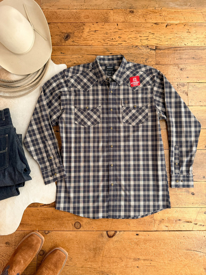 The Evans Ariat Men's Midnight Plaid Button Up from Savannah Sevens western life{&}style is laid flat on a wooden floor, styled with a cowboy hat, dark jeans, brown boots, and next to a white rug.