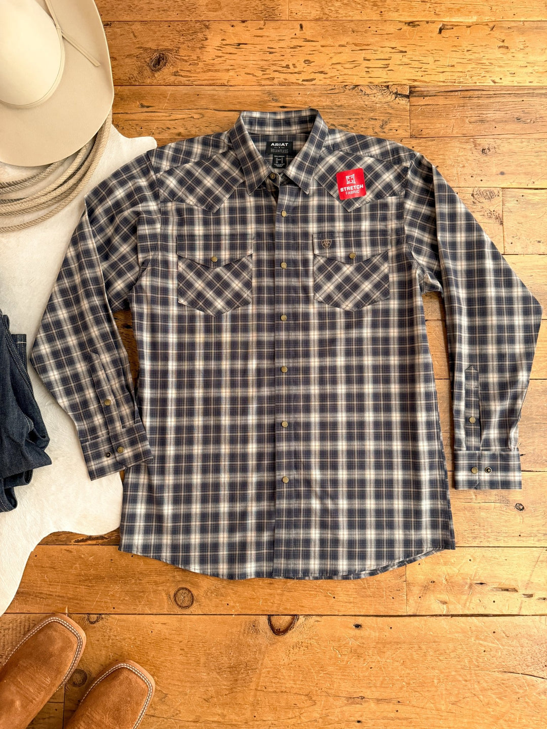 A Savannah Sevens western life{&}style Evans Ariat Men's Midnight Plaid Button Up shirt with two chest pockets is laid flat beside a white cowboy hat, dark jeans, brown boots, and a red Stetson Stretch sticker.