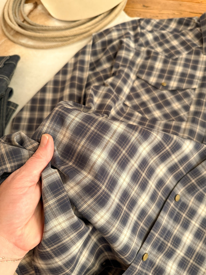 A hand holds the Evans Ariat Men's Midnight Plaid Button Up from Savannah Sevens western life{&}style, highlighting its soft blue and white plaid texture. Coiled rope and another folded shirt can be seen in the background.