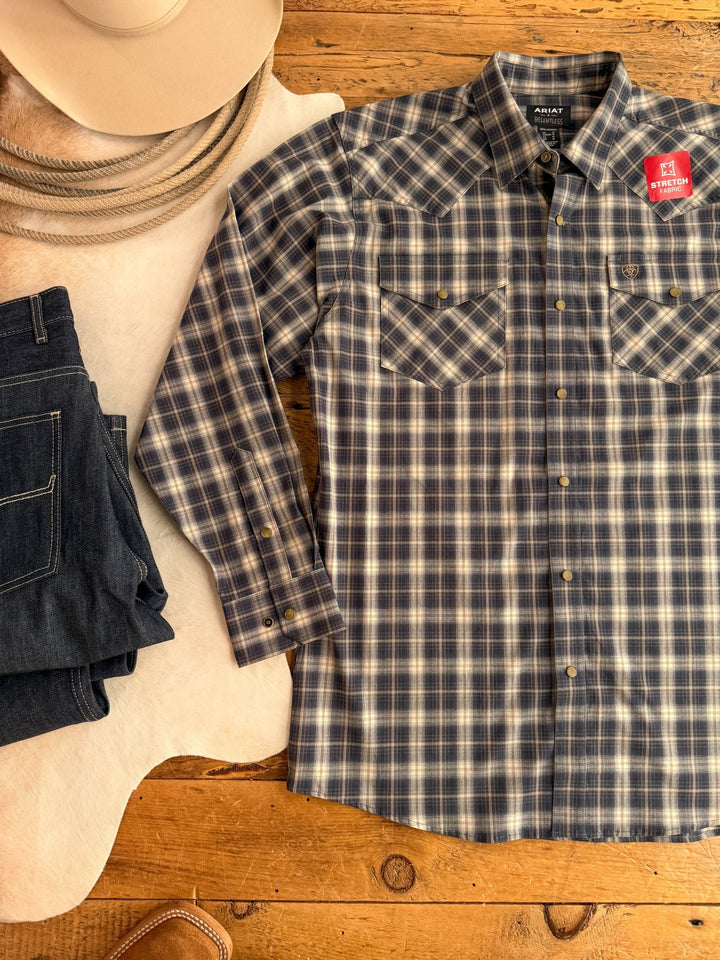 The Evans Ariat Men's Midnight Plaid Button Up by Savannah Sevens western life{&}style, a blue plaid long sleeve shirt with pearl snaps and two chest pockets, is styled beside dark jeans, a cowboy hat, rope, and a cowhide rug on wood. Features moisture wicking fabric.