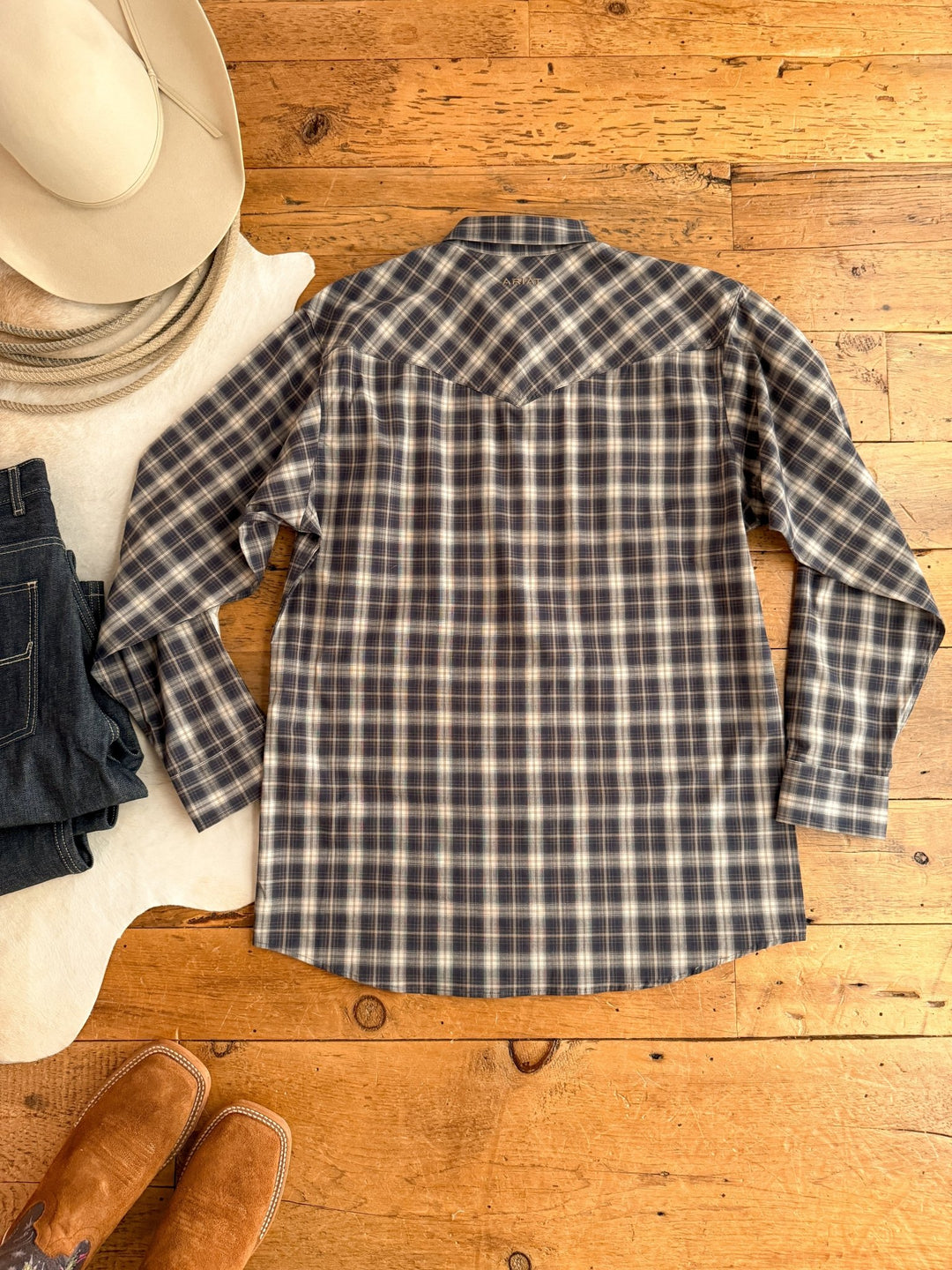 A Savannah Sevens western life{&}style Evans Ariat Men's Midnight Plaid Button Up lies on a wooden floor with a beige cowboy hat, coiled rope, dark jeans, and brown boots. The shirt offers moisture-wicking fabric for all-day comfort.