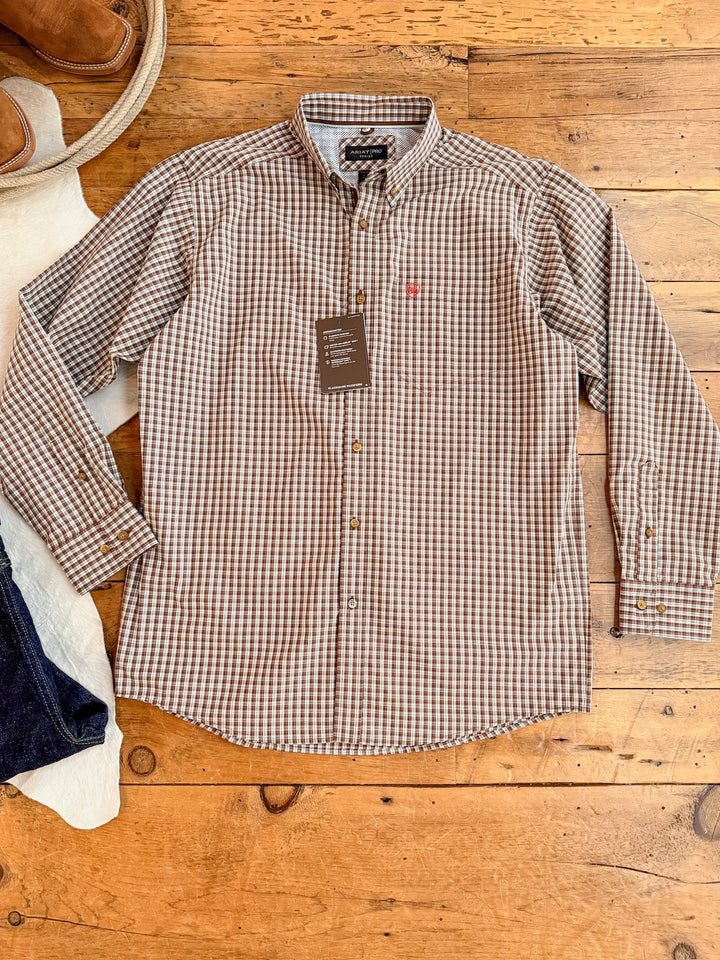 The Everett Ariat Men's Chocolate Plaid Button Up by Savannah Sevens western life{&}style, with brown and white checks and tag attached, is laid flat on a wooden floor beside jeans and tan shoes. This sun protection shirt blends classic style with comfort.
