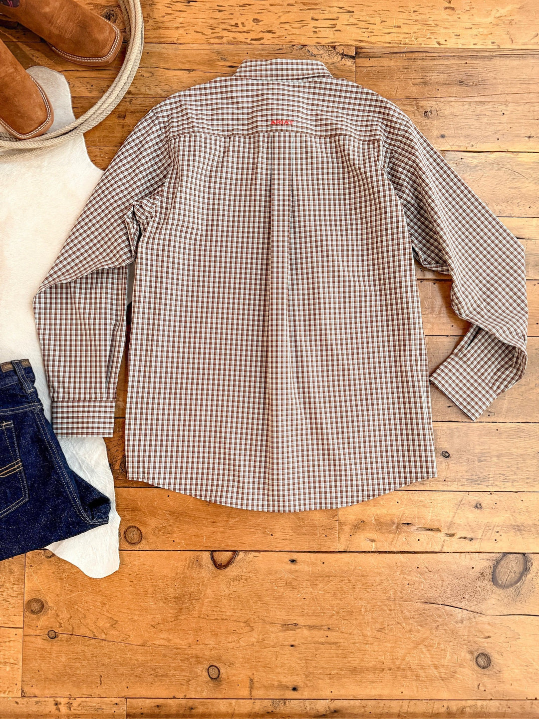 The Everett Ariat Men's Chocolate Plaid Button Up from Savannah Sevens western life{&}style is laid flat on a wooden floor, paired with blue jeans, a white sweater, and brown boots. This long-sleeve offers classic style and outdoor sun protection.