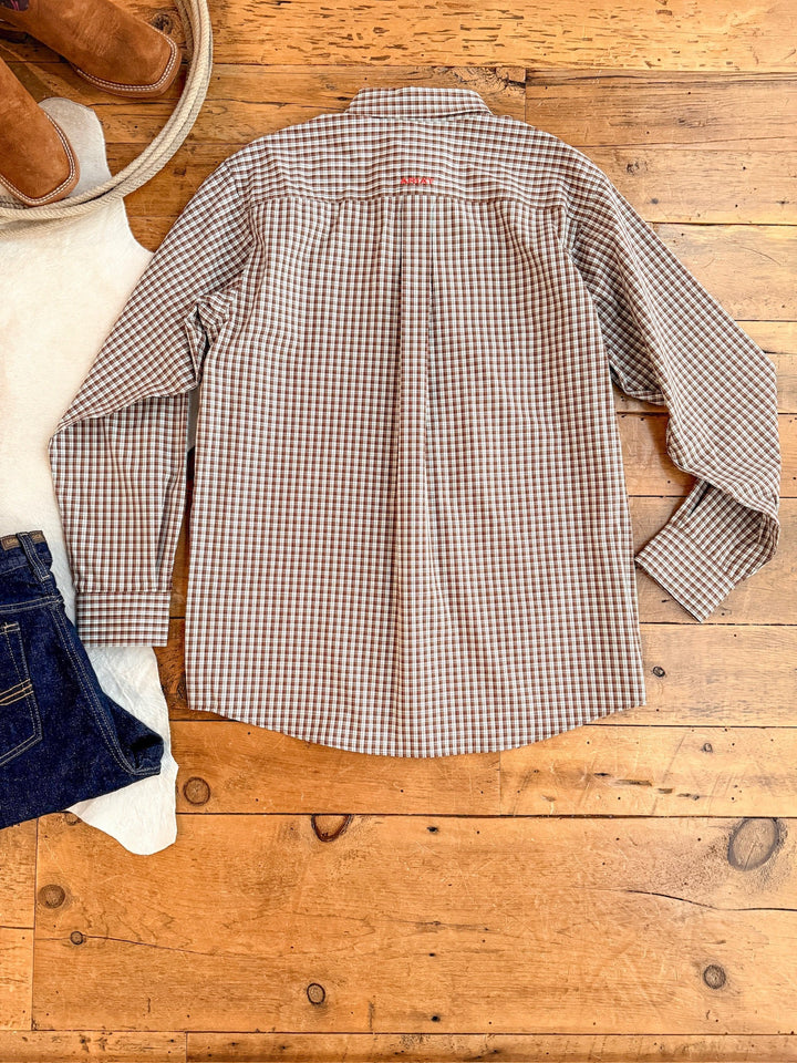 The Everett Ariat Men's Chocolate Plaid Button Up from Savannah Sevens western life{&}style is laid flat on a wooden floor, paired with blue jeans, a white sweater, and brown boots. This long-sleeve offers classic style and outdoor sun protection.