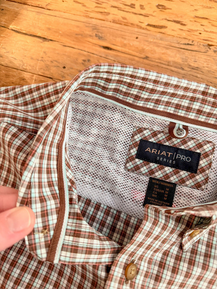 A close-up of the Everett Ariat Men's Chocolate Plaid Button Up by Savannah Sevens western life{&}style shows its brown and white collar, buttons, labels, and inner mesh lining, with a hand holding the collar against a wooden background.