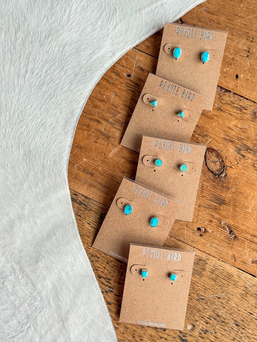 Five pairs of Falcon Earrings by Savannah Sevens western life{&}style are arranged diagonally on brown Peyote Bird cards atop a wooden surface, beside light grey furry fabric.
