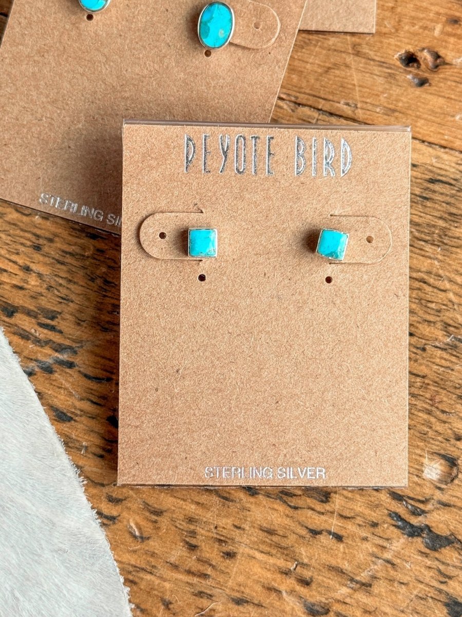 The Falcon Earrings by Savannah Sevens western life{&}style are square Kingman turquoise studs on a brown card labeled STERLING SILVER, displayed on wood with another turquoise jewelry card partially visible.