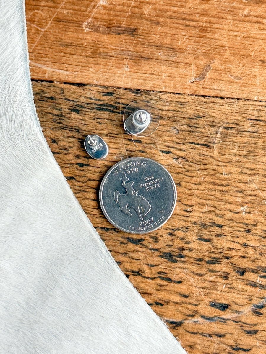 A Wyoming state quarter on wood beside two metal earring backs, part of light-colored cowhide in the corner—ideal for displaying Falcon Earrings by Savannah Sevens western life{&}style.