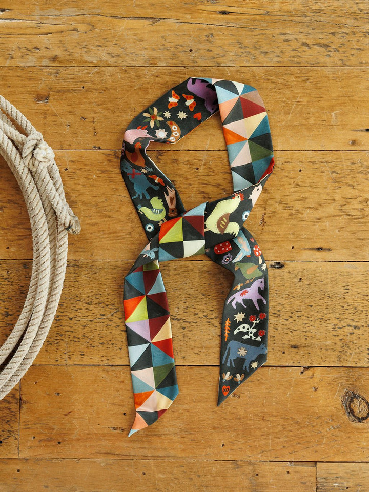 A Farm Folk Silk Twilly by Savannah Sevens western life&style, a long, narrow silk western tie scarf with a geometric print on one side and a whimsical folk-art animal pattern on the other, tied in a knot on a wooden surface next to a rope.