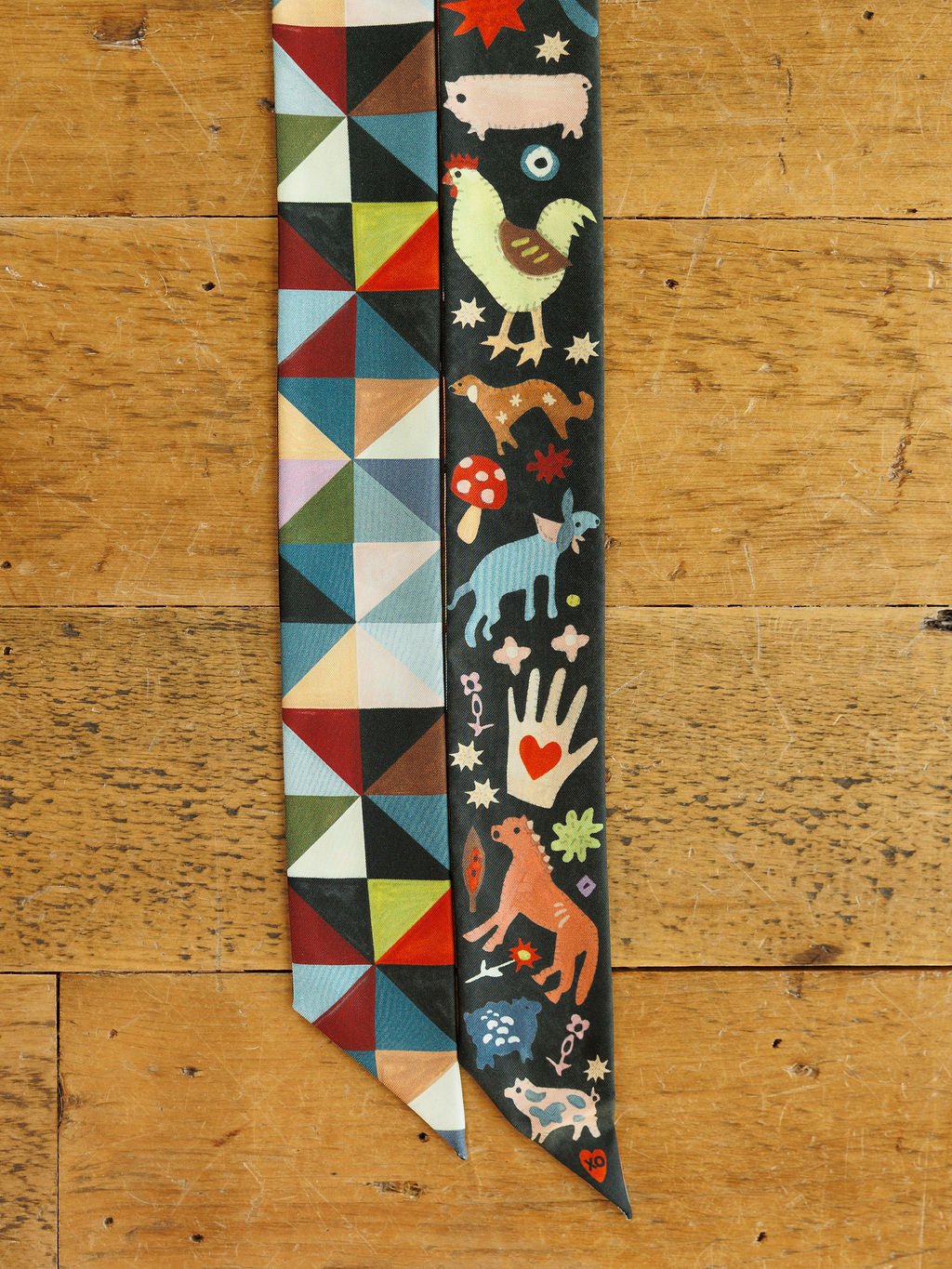Two long, thin Farm Folk Silk Twilly scarves lie side-by-side: one with a multicolored geometric print and the other featuring a dark background with illustrated animals, a heart-adorned hand, and mushrooms.