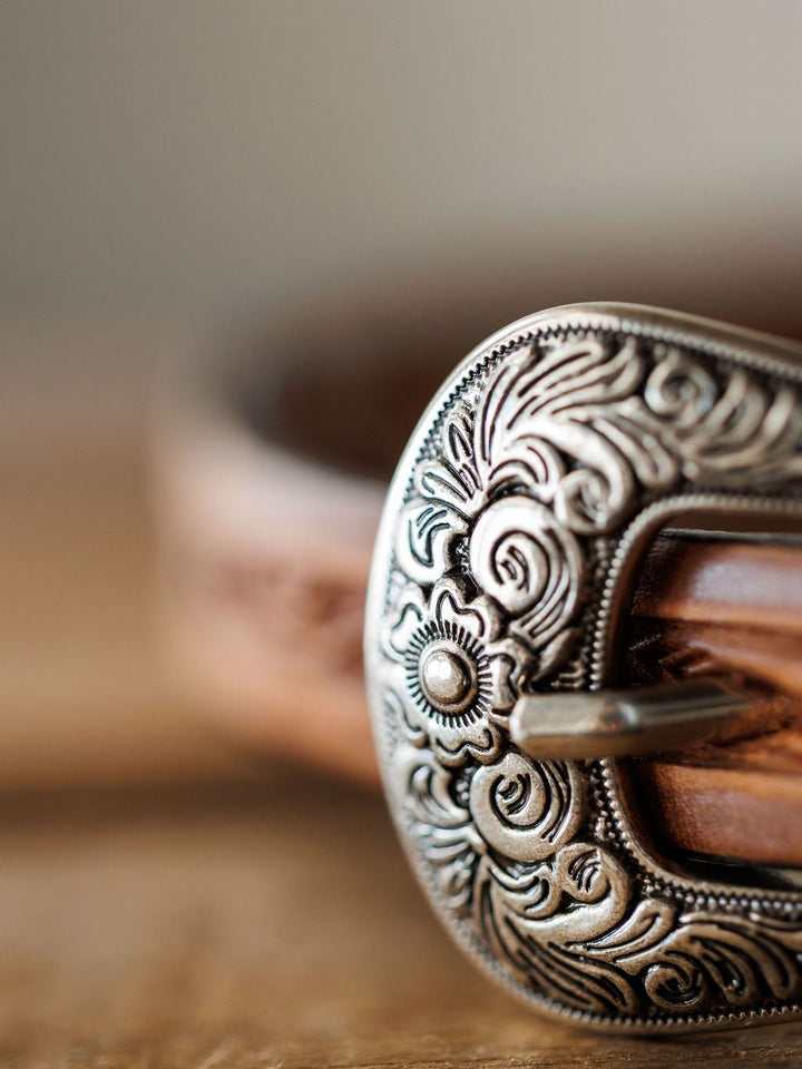 A close-up of the Savannah Sevens western life{&}style Fayetteville Belt shows its ornate silver buckle with intricate floral designs and finely hand-tooled leather, captured with a shallow depth of field to highlight detailed craftsmanship.