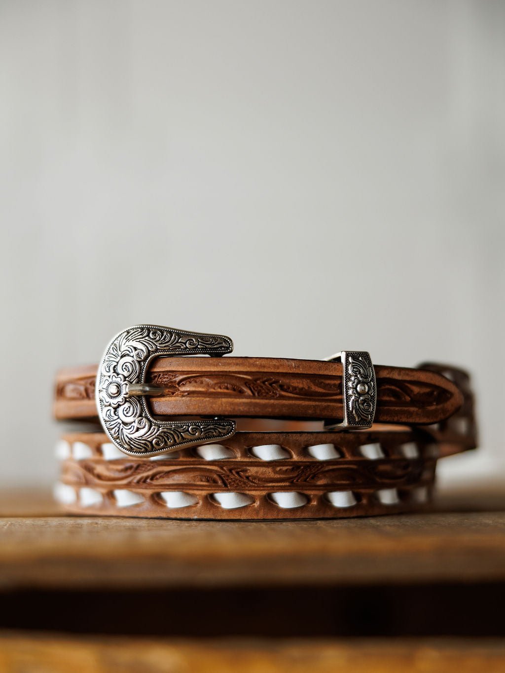 The Fayetteville Belt by Savannah Sevens western life{&}style, featuring intricate hand-tooled leather and an ornate silver buckle, rests on a wooden surface with a neutral backdrop.