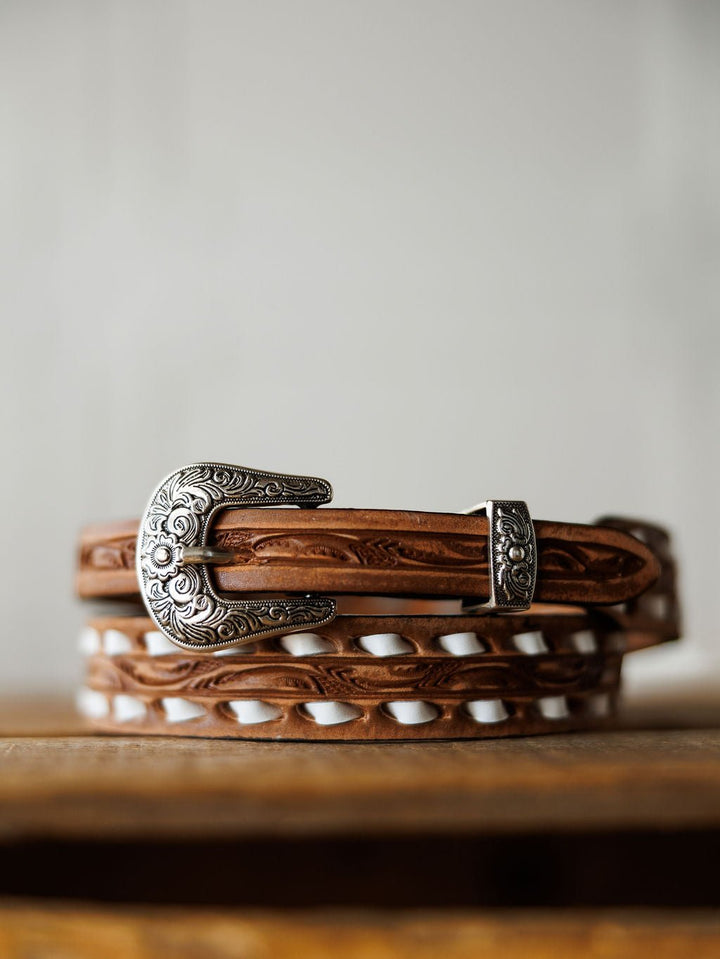 The Fayetteville Belt by Savannah Sevens western life{&}style, featuring intricate hand-tooled leather and an ornate silver buckle, rests on a wooden surface with a neutral backdrop.
