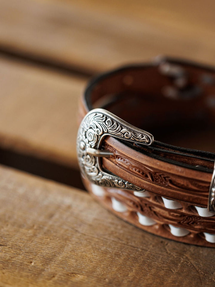 A close-up of the Savannah Sevens western life{&}style Fayetteville Belt, featuring hand-tooled leather, a decorative silver buckle with white accents, and detailed engraving, resting on a wooden surface.
