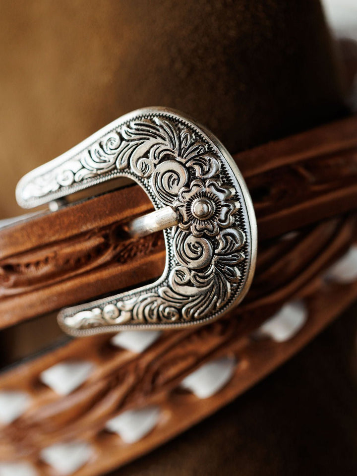 Close-up of an ornate silver belt buckle with intricate floral engravings on the Fayetteville Belt by Savannah Sevens western life{&}style, set against a blurred brown background.