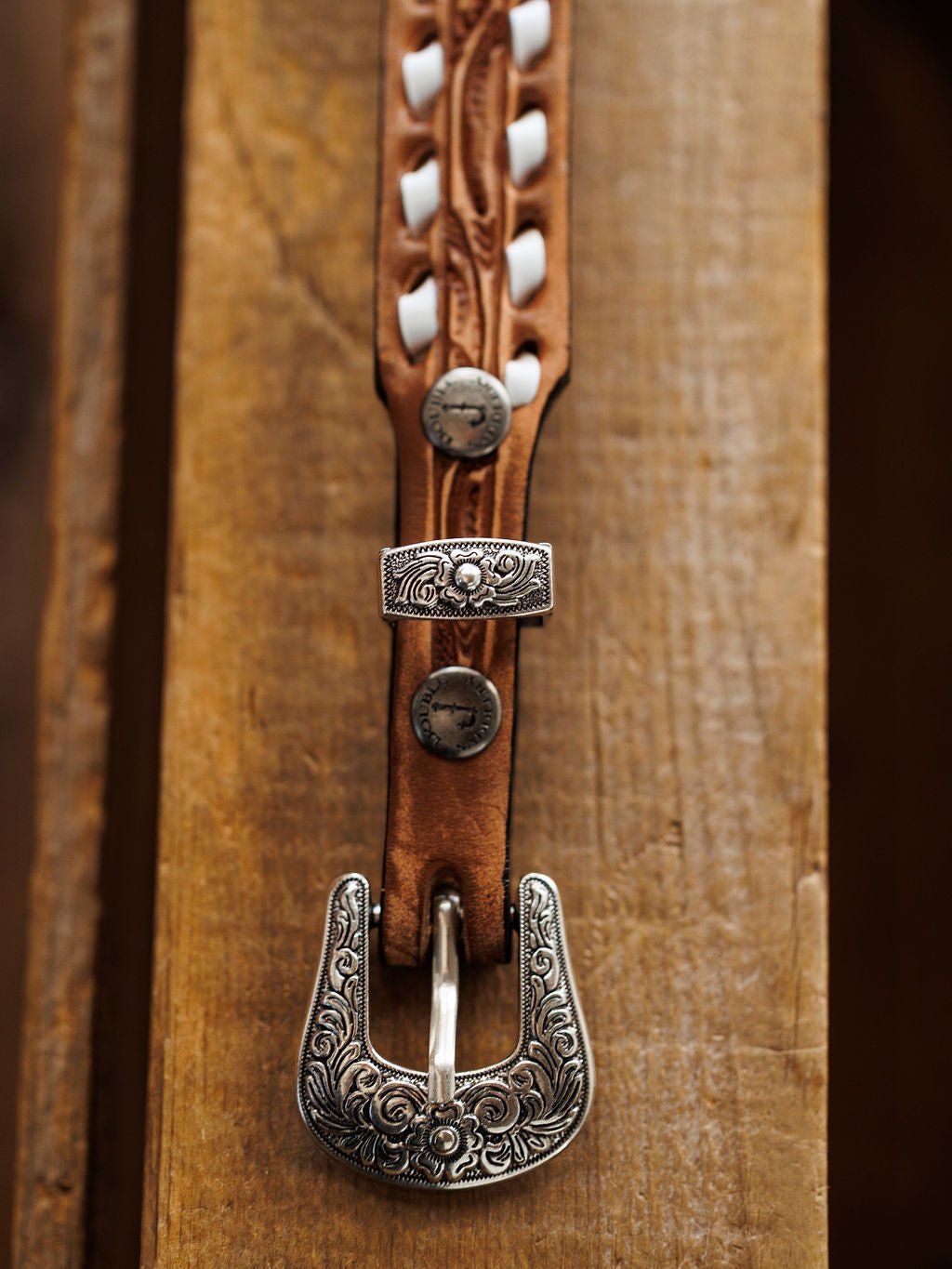 A close-up of the Fayetteville Belt by Savannah Sevens western life{&}style, featuring intricate white stitching and engraved silver hardware on the decorative buckle, displayed on a wooden surface.