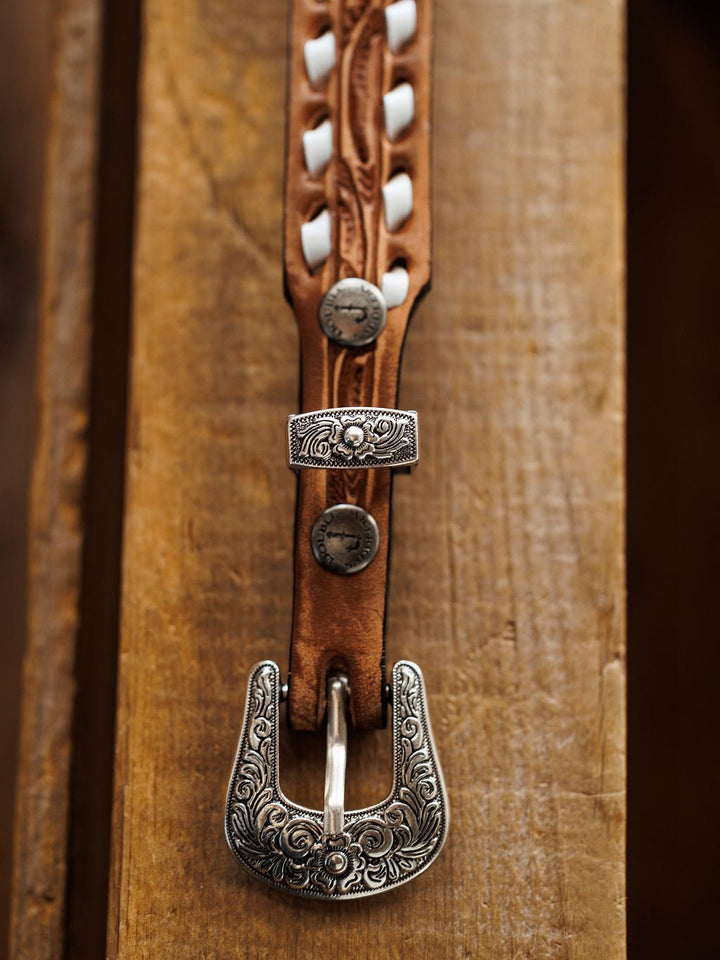 A close-up of the Fayetteville Belt by Savannah Sevens western life{&}style, featuring intricate white stitching and engraved silver hardware on the decorative buckle, displayed on a wooden surface.
