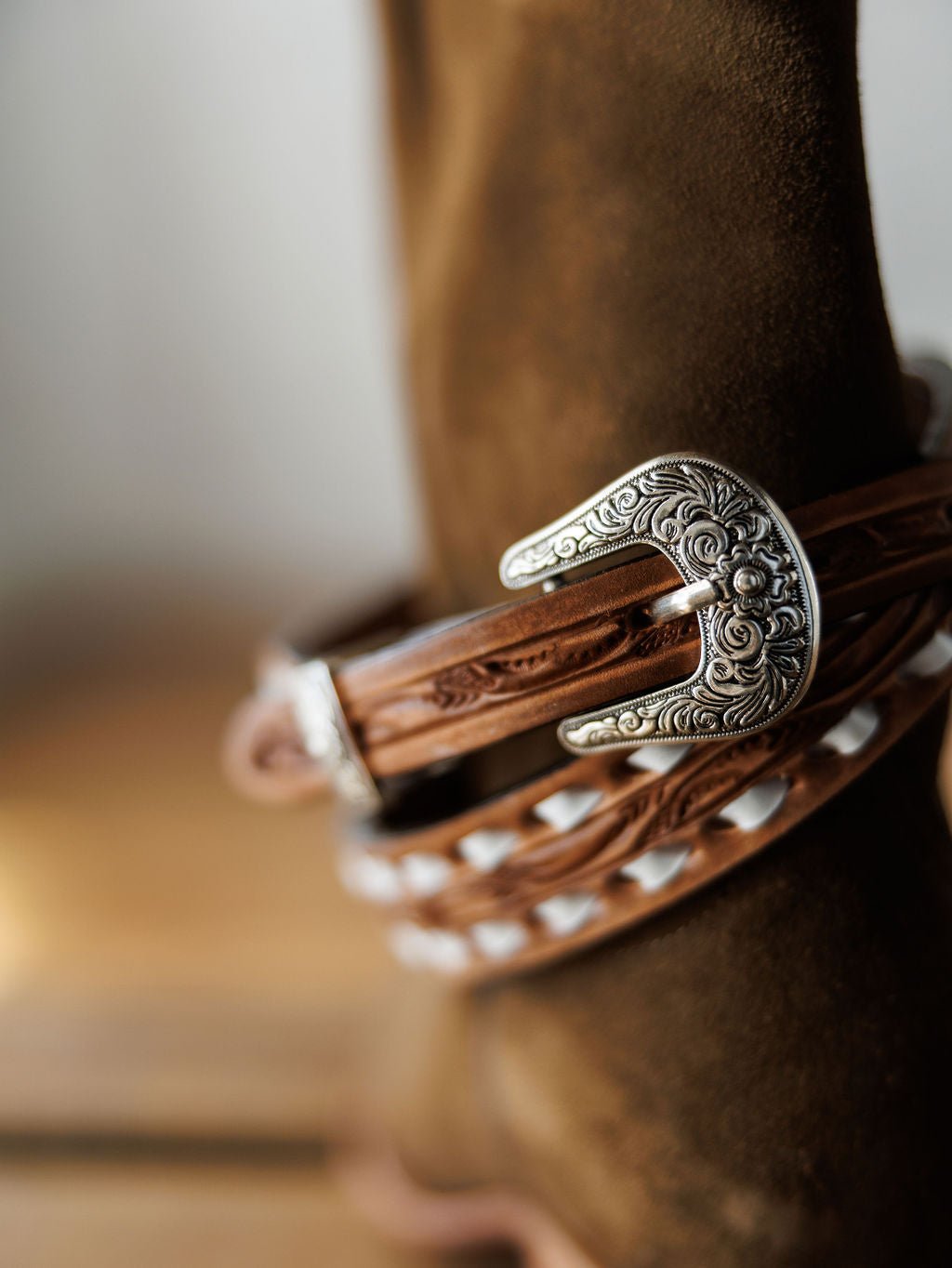 Close-up of a Savannah Sevens western life{&}style Fayetteville Belt’s silver buckle, featuring an intricate floral design, on a hand-tooled leather belt wrapped around a brown suede boot.