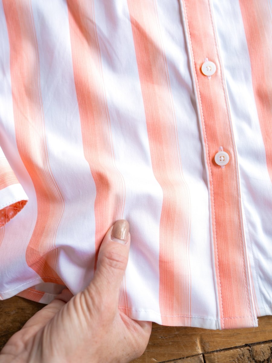 A hand with painted nails holds the hem of The Forada, a wrinkle-resistant striped button-up top by Savannah Sevens western life{&}style, with vertical orange stripes and buttons, laying flat on a wooden surface.